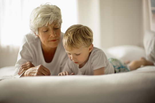Mature woman and grandson lie on their fronts on a bed side by side while looking down.