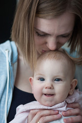 Mom holds a baby in her arms and kisses her on the top of her head. The baby is three months old and she is smiling at the frame