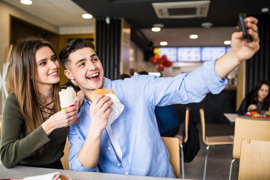 Happy beauty couple eating burger and doner taking selfie together in fast food