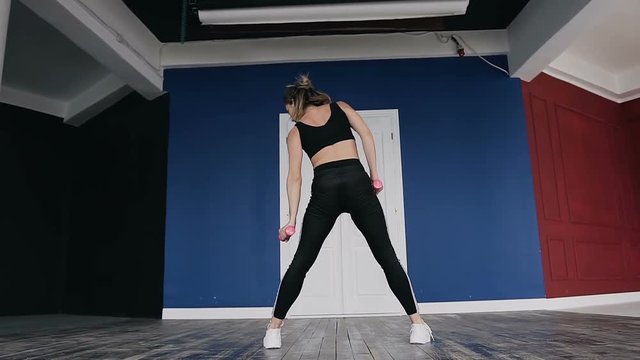 Bending to the side and forward. Rear view of young beautiful blonde girl in sportswear doing bending to the side and forward holding dumbbells in their hands at gym indoors