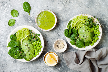 Obraz premium Vegan, detox Buddha bowl with avocado, spinach, micro greens, edamame beans, zucchini noodles and herb green dressing. Top view, grey concrete background