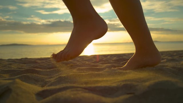 SLOW MOTION, CLOSE UP, LENS FLARE: Unknown female jogger runs barefoot on dry sand by the tranquil ocean. Playful traveler runs along the sunlit sandy beach on calm summer evening. Picturesque seaside