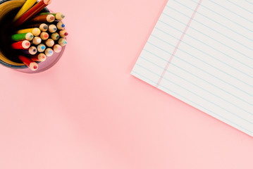 A cup with colored pencils and a white notepad on a flat pink surface