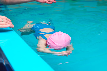 A little girl of European appearance in a pink rubber cap learning to swim in the pool in glasses for swimming