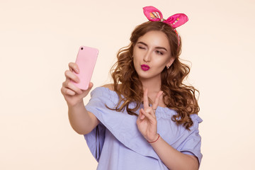 Beautiful young woman in an Pin-up image doing selfie on the phone and showing thumbs up sign of peace. On his head is a pink bandage, ears.