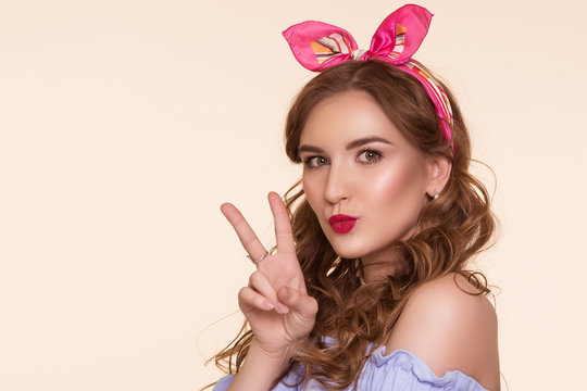 Beautiful young woman in pin-up image and bow on head, on beige background. A sign of peace, a gesture with your fingers.