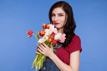 Obraz na płótnie Canvas Beautiful woman, brunette, stands on blue background, in denim overalls and T-shirt with bouquet of tulips. The feminist. International Women's Day, March 8.