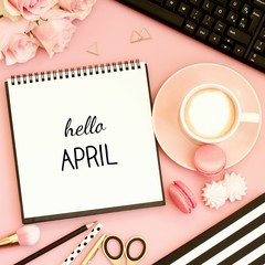 Hello April text on notebook. Office desk table with cup of coffee, sweet snacks, computer and pink...