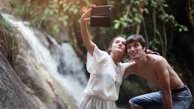 Young happy couple taking selfie outside by waterfall with mobile phone in tropical jungle. 1920x1080