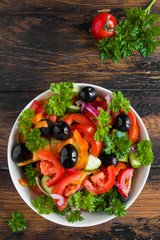 Vegetable salad with fresh tomatoes, cucumbers, black olives, red onion and parsley in a white bowl on the wooden brown table, top view.