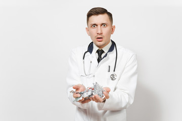 Sad upset shocked handsome young doctor man isolated on white background. Male doctor in medical uniform, stethoscope holding packing tablets, pills. Healthcare personnel, health, medicine concept.