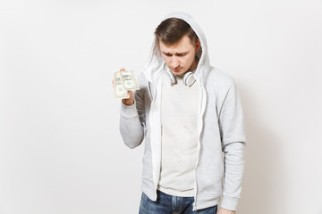 Young handsome man student in t-shirt, blue jeans and light sweatshirt with hood with headphones holds two one-dollar bills and sadly looks down isolated on white background. Concept of emotions