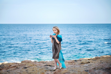young blond girl in a fairy-tale dress like a bird holding a royal crown strolling along the shore