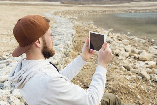 Hipster with a beard in a cap and a sweatshirt with a tablet in his hands takes pictures on a tablet while being on the nature by the lake