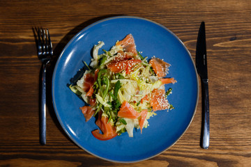 A horizontal image of a bright blue plate with salad, containing salmon, salad leaves, arugula, tomatoes, sesame seeds