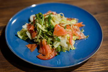A horizontal image of a bright blue plate with salad, containing salmon, salad leaves, arugula, tomatoes, sesame seeds