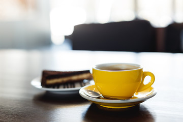 A horizontal image of a yellow cup of black coffee and a plate with a piece of chocolate cake. Bokeh background.