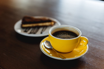 A horizontal image of a yellow cup of black coffee and a plate with a piece of chocolate cake. Bokeh background.