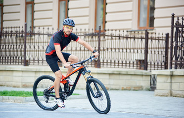 Bicyclist in professional cycling garment and protective helmet looking to camera, riding bike near beautiful buildings. Man training, improving hobby, getting ready for contest