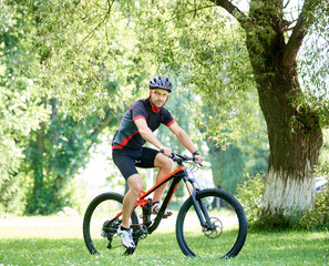 Fototapeta na wymiar Sporty male biker in cycling sportswear and protective gear riding bike along green park alleys, smiling and looking at camera. Concept of healthy lifestyle, connecting with nature