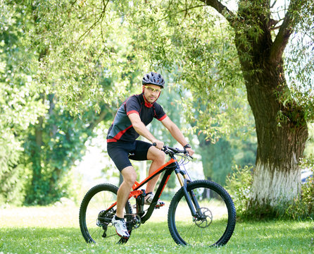 Sporty male biker in cycling sportswear and protective gear riding bike along green park alleys, smiling and looking at camera. Concept of healthy lifestyle, connecting with nature