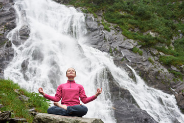 Fototapeta na wymiar Connecting with nature. Young beautiful female practising yoga on stone in front of beautiful Balea waterfall in Romania. Woman yogini green rock amazing breathtaking scenery view nature happy calm