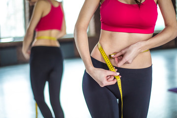 Athletic smiling slim woman measuring her waist by measure tape after workout in gym