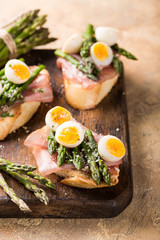 Fototapeta na wymiar Easter breakfast or lunch with sendwich with ham, asparagus and quail eggs on old wooden chopping board. Spring food concept with copy space.