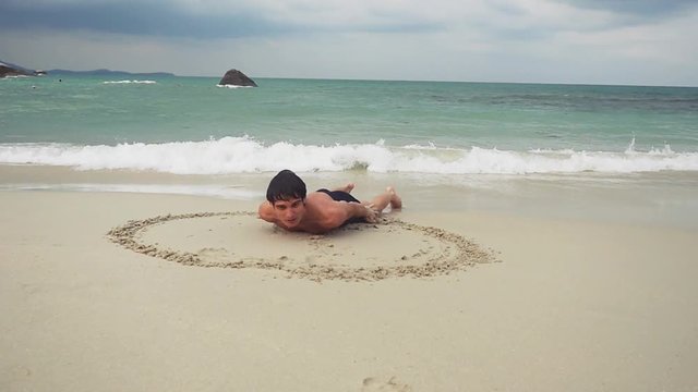 Young man lies on the sand draws a heart symbol on the wet sand on the seashore. slow motion. 1920x1080