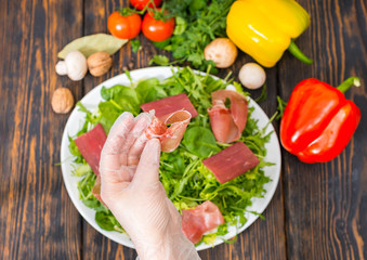 High angle view of vegetables, hands in glove holding jamon above white plate with dried meat and mix of lettuce leaves
