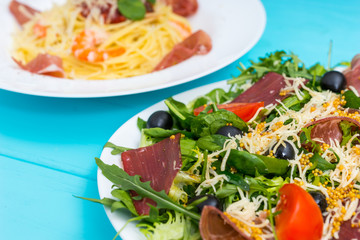 Close up of delicious salad in white plate on blue wooden table, spaghetti in the background