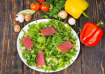 High angle view of tomatoes, red and yellow pepper, dill and parsley around white plate with dried meat and mix of lettuce leaves