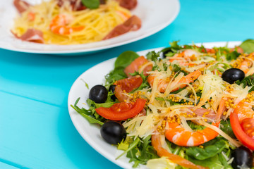 Close up of delicious salad in white plate on blue wooden table, spaghetti in the background