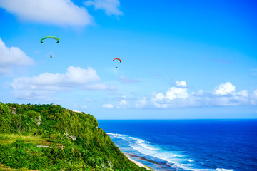 Paragliding over the ocean on the island of Bali. The blue sky shimmers with the blue ocean. The parasutists fly high above the precipice like birds. Aerial view with copy space
