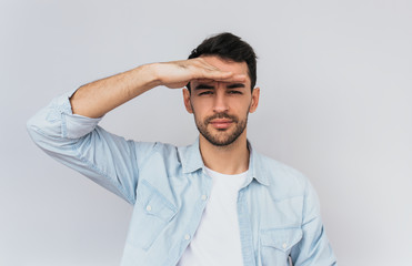 Studio portrait of handsome positive male keeping his hand over his eyes to protect from the sunrays. Bearded man looking at the camera keep his hand on forehead on studio wall background. People