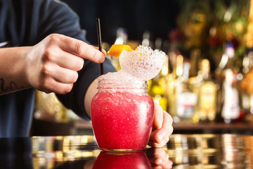 Bartender is decorating red cocktail in jar with frappe at bar counter background.