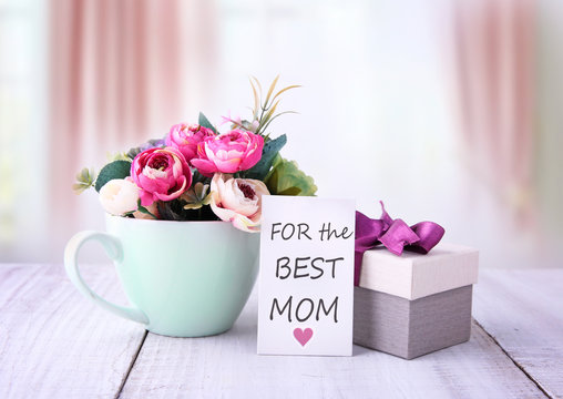Happy mothers day gifts with red white roses in a cup, and gift tag with text „For the best mom“, presented on wooden table with bright backround