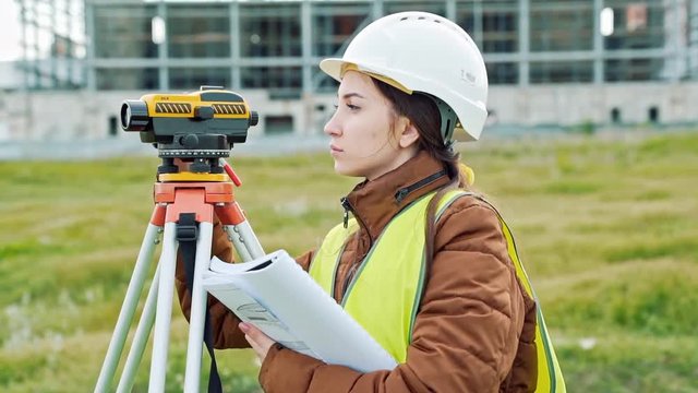A young woman surveyor in work clothes and helmet adjusts the equipment and produces calculations on the construction site. The concept of landscape design, geodesy