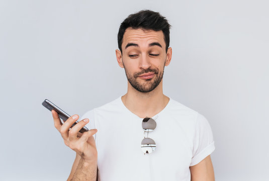 Studio Portrait Of Annoyed Young Caucasian Bearded Man In Shades And Tank Top White Tshirt Getting Disgusted Because Of His Mobile Phone Battery Running Down Or Loud Conversation. People, Technology