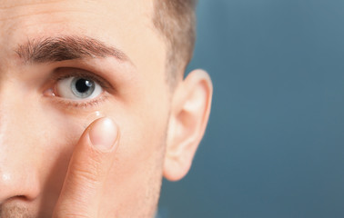 Young man putting contact lens in his eye on color background