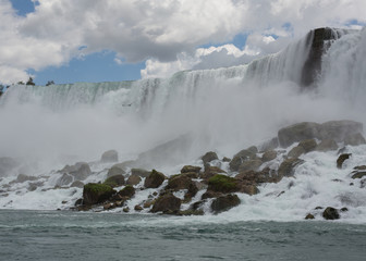 Niagara falls seen from the bottom with puffy white clouds in the sky that match the white foam of the falls. 