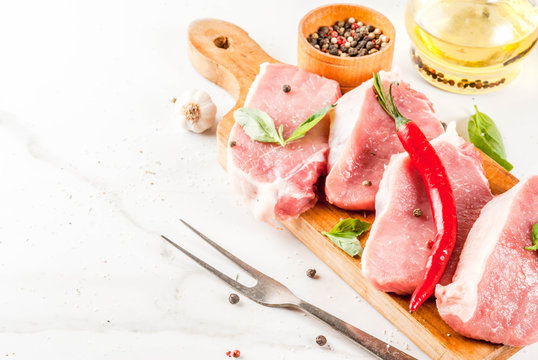 Raw meat, pork steaks with spices, herbs, olive oil, white marble background on cutting board, copy space