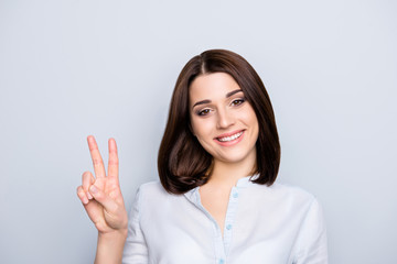 Portrait of modern, smiling, nice girl with short hairstyle showing v-sign, two fingers, looking to the camera, isolated on grey background