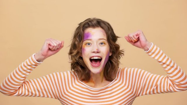 Portrait of emotional woman 20s with colorful designer painting on face shouting and clenching fists like lucky person slow motion, isolated over beige background. Concept of emotions