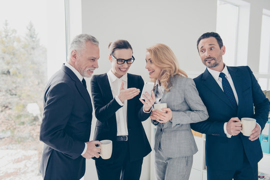 Four attractive, cheerful, stylish business persons using smart phone, watching funny, comic video, photo, drinking coffe, tea, enjoying leisure, standing in workplace, workstation