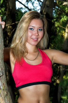 blonde in sportswear in a thin pink t-shirt and black tight-fitting shorts posing in the Park in the summer in green bushes illuminated by the sun