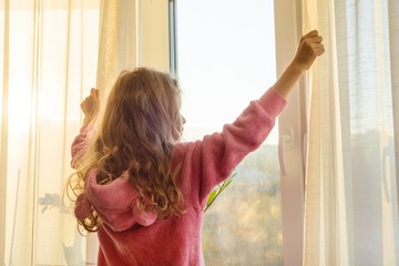 Fototapeta na wymiar Good morning. Girl child in pajamas opens curtains and looks out the window.