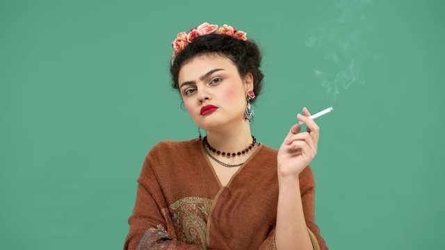 Portrait of elegant woman with thick eyebrows as Frida Kahlo with roses in hair looking seriously while smoking cigarette, isolated over green background. Stylization concept
