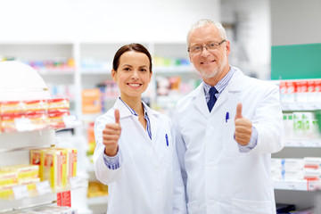 medicine, healthcare and technology concept - happy smiling apothecaries at pharmacy showing thumbs...