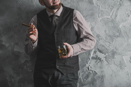 Man in vest standing, leaning against the wall, holding a glass of whiskey. No face